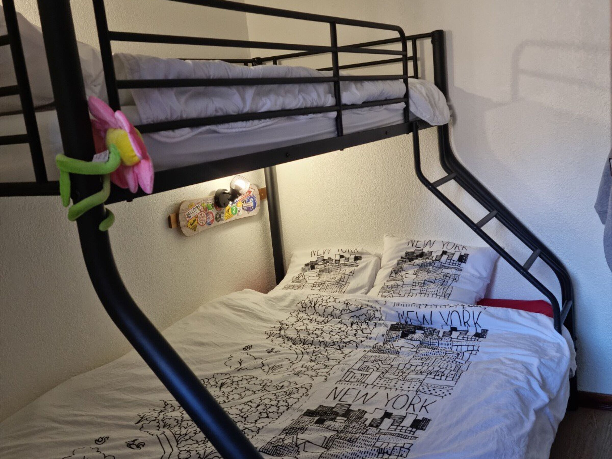 Un lit superposé avec une couette et des oreillers décorés sur le thème d’un paysage urbain, un jouet vert et un objet fixé au mur, orné d’autocollants.