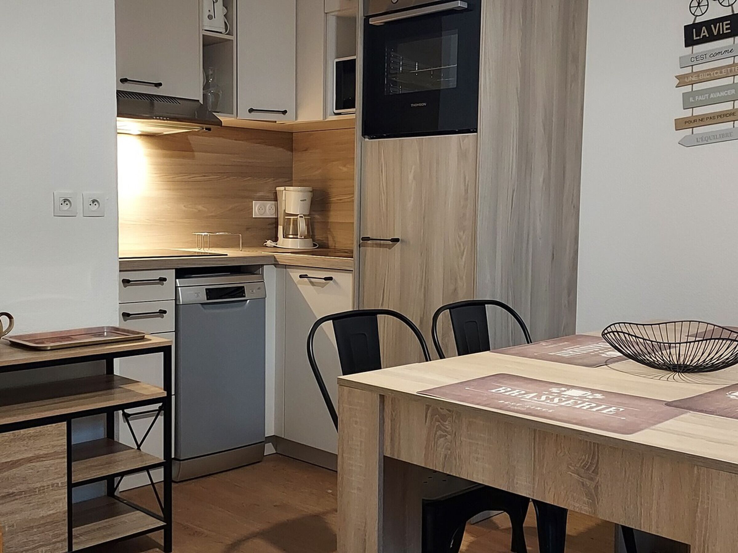 A compact kitchen with a dishwasher, a coffee maker, and a microwave. There is a dining table with chairs and a decorative bowl.
