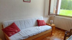 Una habitación pequeña y acogedora con un sofá tipo banco de madera, dos almohadas rojas, una mesita de noche con lámpara y una ventana con vistas a zonas verdes.