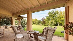 Eine Terrase mit Korbmöbeln, einem Tisch und Blick auf einen gepflegten Rasen und Bäume.
