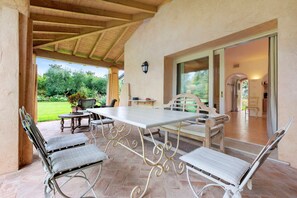 An outdoor patio with a table and chairs, a view of the garden, and a covered area with a door leading inside.