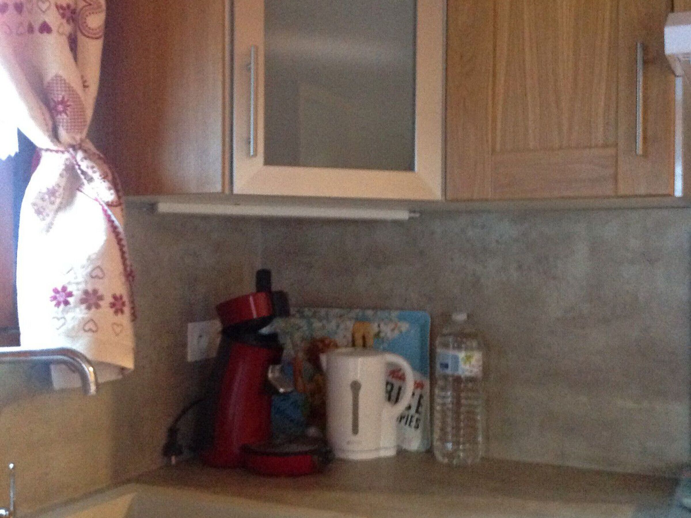 Une cuisine avec des meubles de rangement en bois, une machine à café rouge, une tasse blanche et une bouteille d’eau posée sur le comptoir.