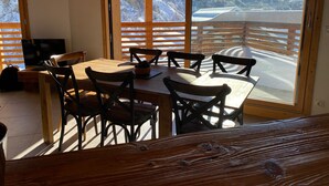 A dining area with a wooden table and chairs, a television, and a view of snow-covered mountains through large windows. - Chalet for 8 facing south, terrace, private parking (Valloire)