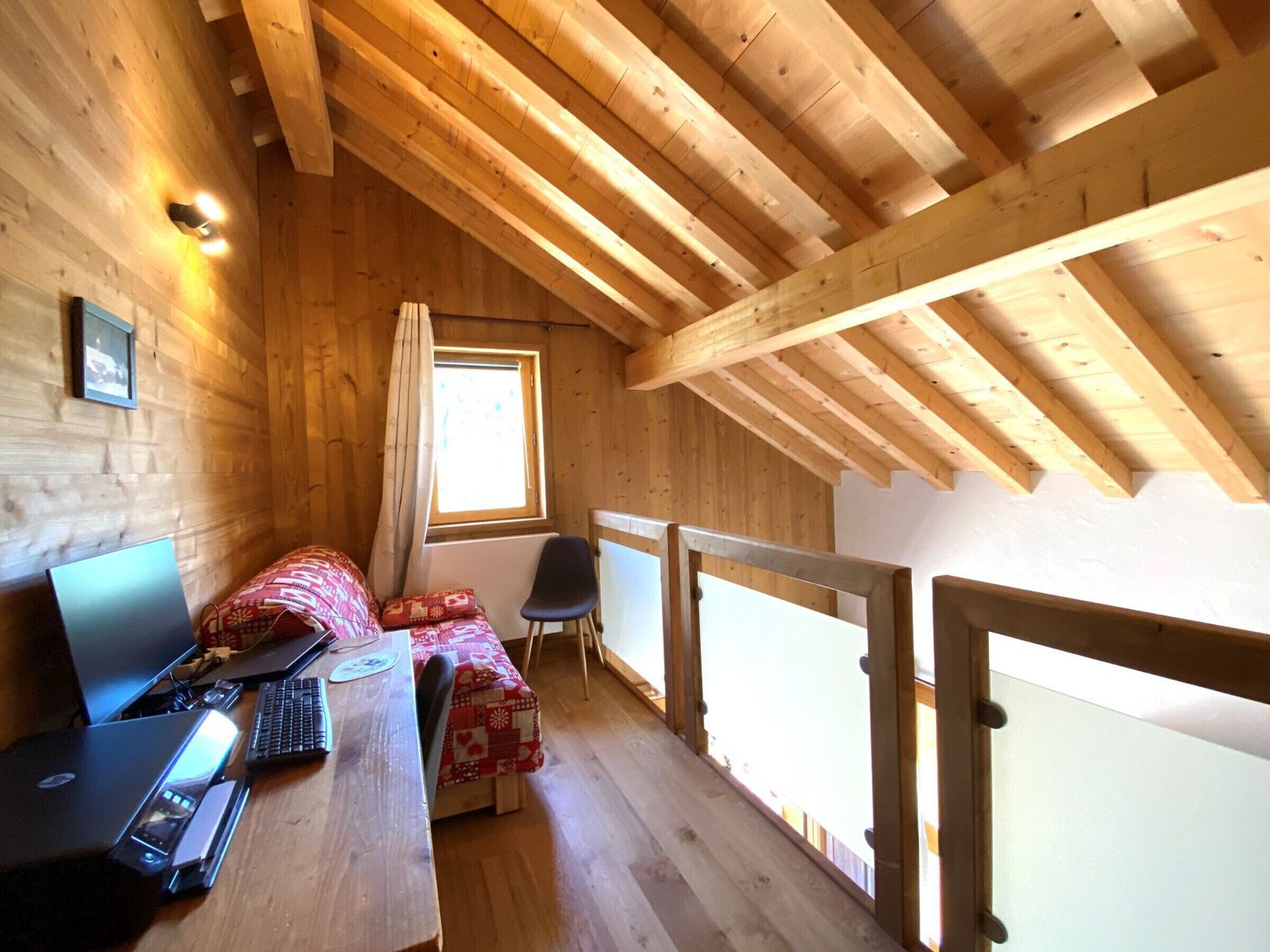 Een knus zolderkamer met houten balken, een bureau met een laptop, een klein bed en een raam met gordijnen.