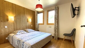 Ein Schlafzimmer mit einer Holzwand, einem Bett mit gemusterten Betttextilien, zwei Nachttischlampen, einem Sessel und einem an der Wand montierten Fernseher.