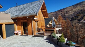 Eine Holzhütte mit einer Steinterrasse, einer überdachten Sitzgelegenheit im Freien und Blick auf die umliegenden Berge.