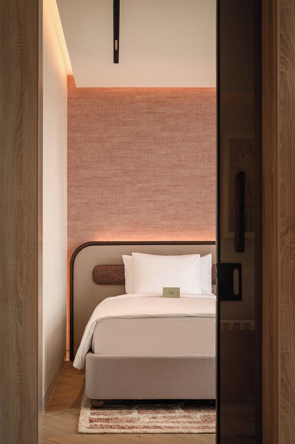 A modern hotel room with a minimalist design, featuring a bed with white linens, a wooden headboard, and a small bedside table.