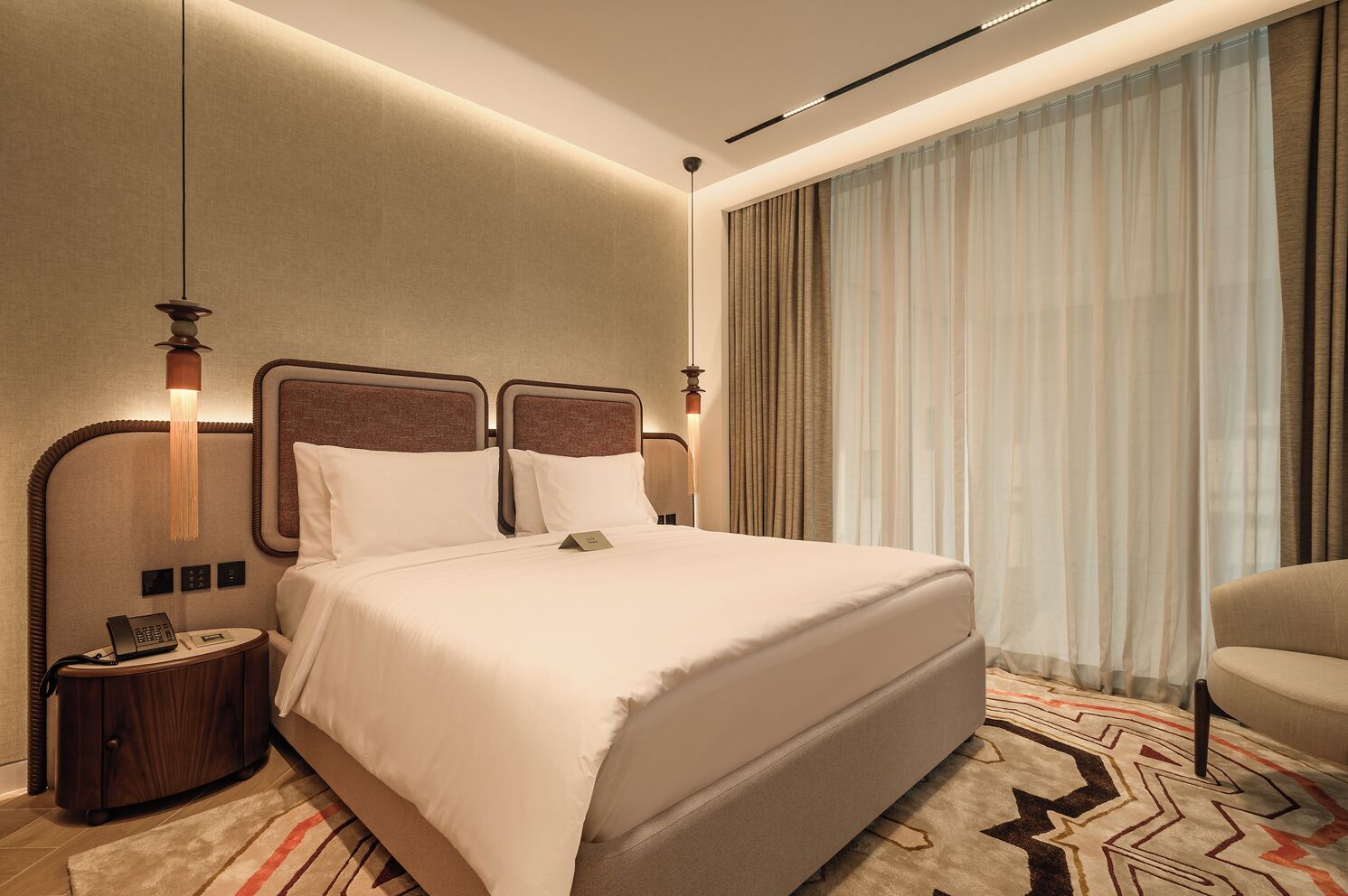 A hotel room with a large bed, two bedside tables, a lamp, a telephone, and a view of the outdoors through sheer curtains.