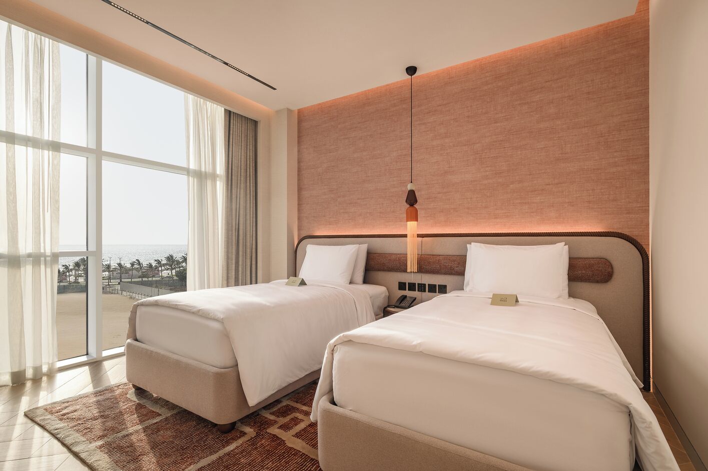 A hotel room with two beds, a large window with a view of the beach, and a modern light fixture above the beds.