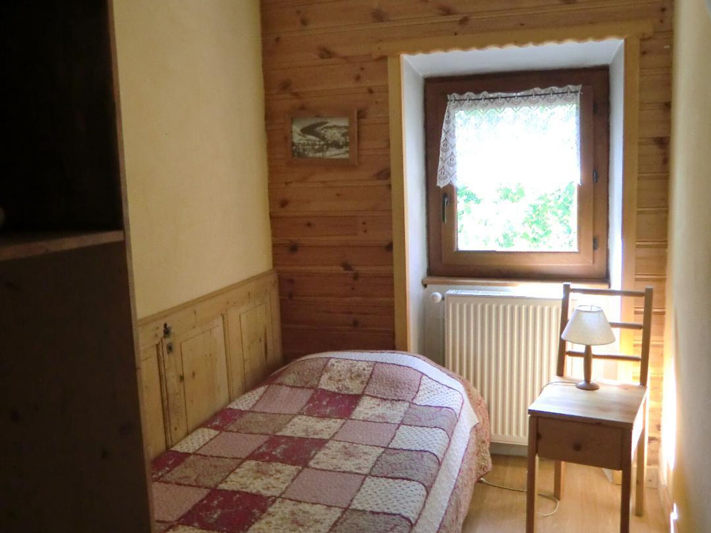 Een slaapkamer met een houten wand, een raam met gordijn, een radiator, een bed met ruitjesdeken en een klein tafeltje met lamp.
