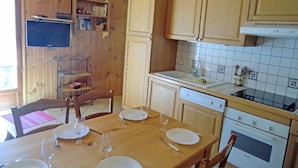 Een houten interieur met een eettafel voor vier personen, een keuken met witte kasten en een betegelde spatwand, en een aan de muur bevestigde televisie.
