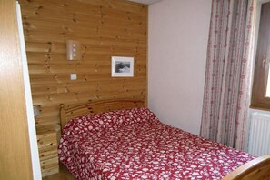A room with a wooden headboard, a bed with a red floral bedspread, a framed picture on the wall, and a window with patterned curtains.