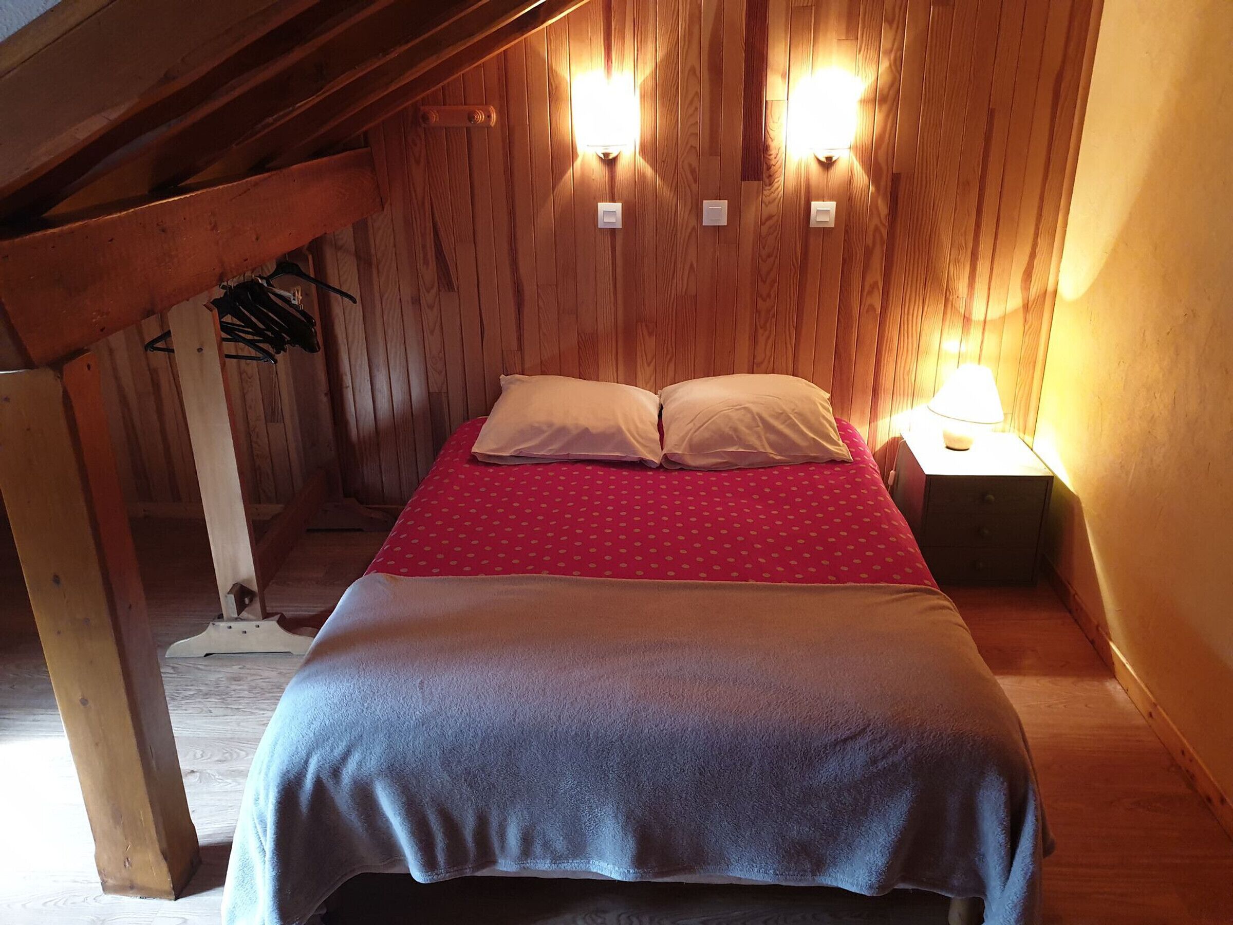 A cozy wooden room with a bed, two wall-mounted lights, and a nightstand with a lamp.