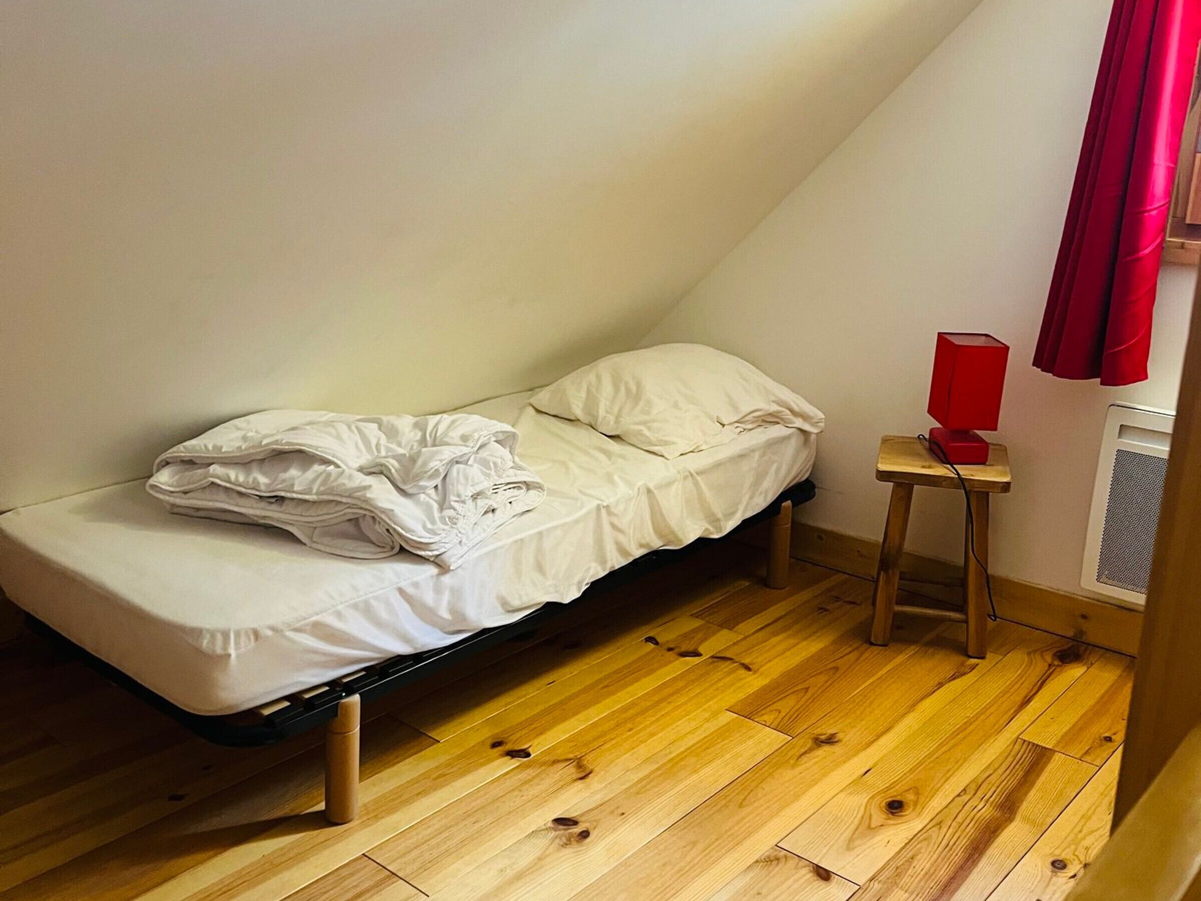 A small, single-bed room with a sloped ceiling, a wooden floor, a red lamp on a small table, and a window with red curtains.