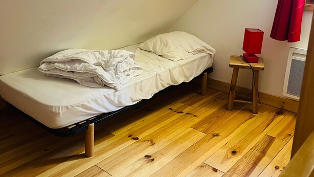 Une petite chambre avec un lit simple, un plafond en pente, un plancher en bois, une lampe rouge posée sur une petite table et une fenêtre ornée de rideaux rouges.