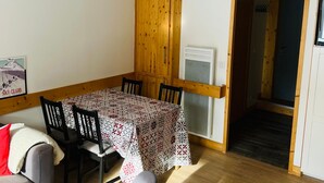 A dining area with a table covered in a patterned tablecloth, surrounded by chairs. A wooden cabinet is mounted on the wall, and a door leads to another room. - Triplex 3 rooms with loft, 6 people, terrace, private parking (Valloire)