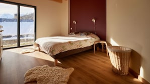 A bedroom with a large window, wooden beams, a bed, a chair, and a wicker basket. - Spacious chalet for 11, 4 rooms, parking, sunny terrace (Valloire)