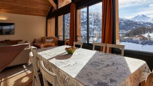 A cozy room with a wooden ceiling, large windows with curtains, a dining table with a floral tablecloth, and a view of snow-covered mountains. - Spacious chalet for 11, 4 rooms, parking, sunny terrace (Valloire)