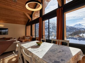 A cozy room with a wooden ceiling, large windows with curtains, a dining table with a floral tablecloth, and a view of snow-covered mountains. - Spacious chalet for 11, 4 rooms, parking, sunny terrace (Valloire)