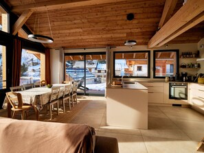 A modern kitchen with a central island, wooden beams on the ceiling, and large windows offering a view of the outside. - Spacious chalet for 11, 4 rooms, parking, sunny terrace (Valloire)
