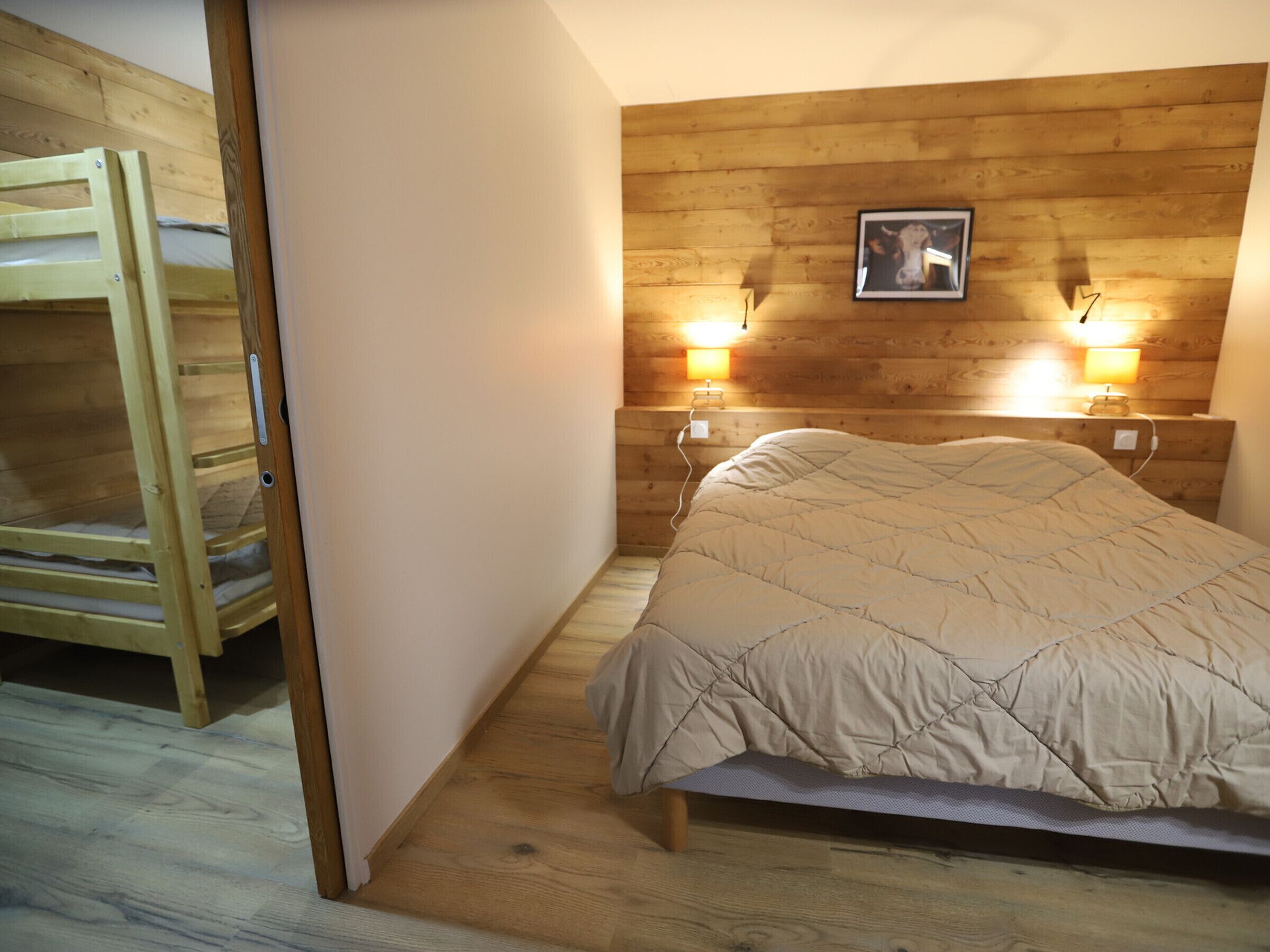 Une chambre avec un lit superposé en bois, un lit simple avec une couverture matelassée, deux lampes de chevet et un tableau encadré au mur.