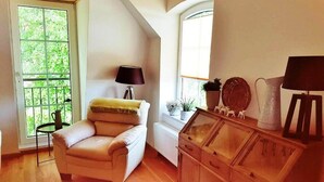 Une pièce avec un plancher en bois, un fauteuil beige, une lampe, un radiateur, un meuble en bois et une fenêtre donnant sur un paysage verdoyant.
