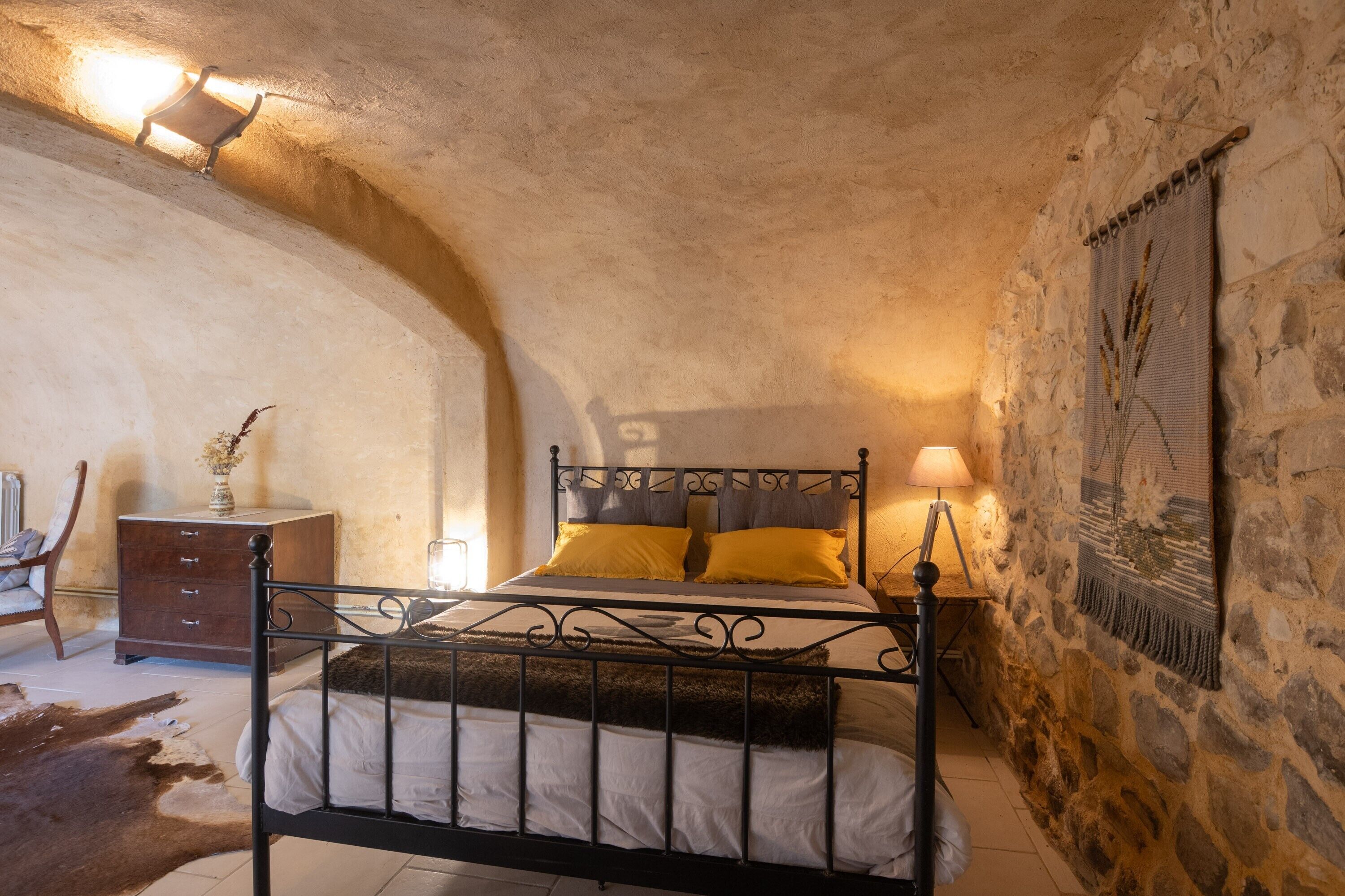 A bedroom with a stone wall, a bed with a metal frame, a wooden nightstand, a lamp, and a cowhide rug.