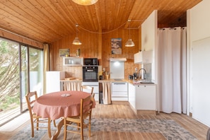A wooden interior with a dining table, chairs, a kitchen area with appliances, and a large window.