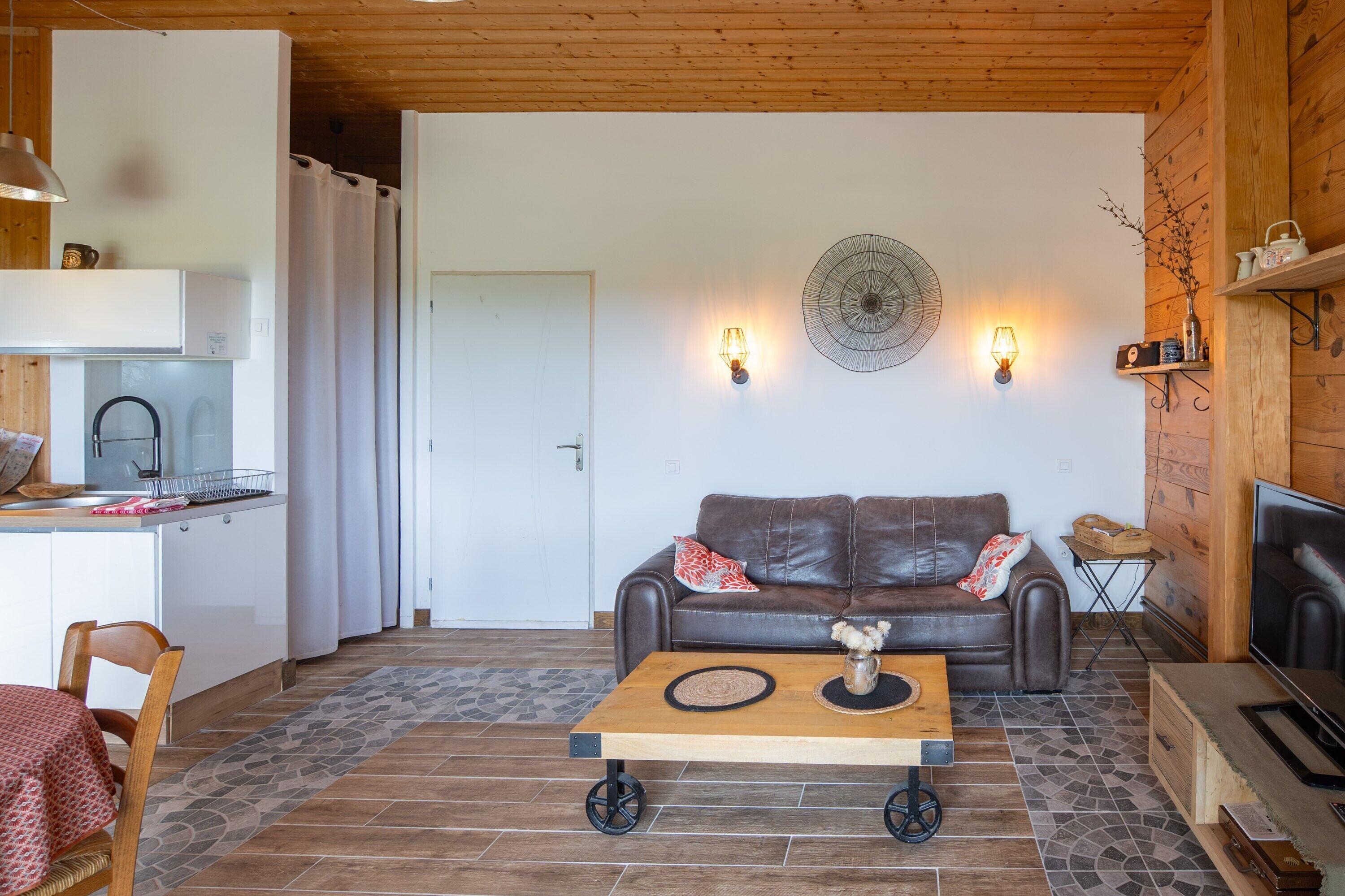 A living room with a brown leather sofa, a wooden coffee table, a TV, and a kitchen area with a fridge and a sink.