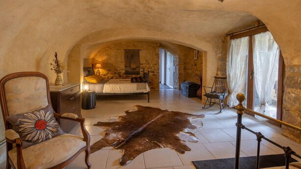 A rustic bedroom with a stone wall, a bed, a chair, a cowhide rug, and a chandelier.