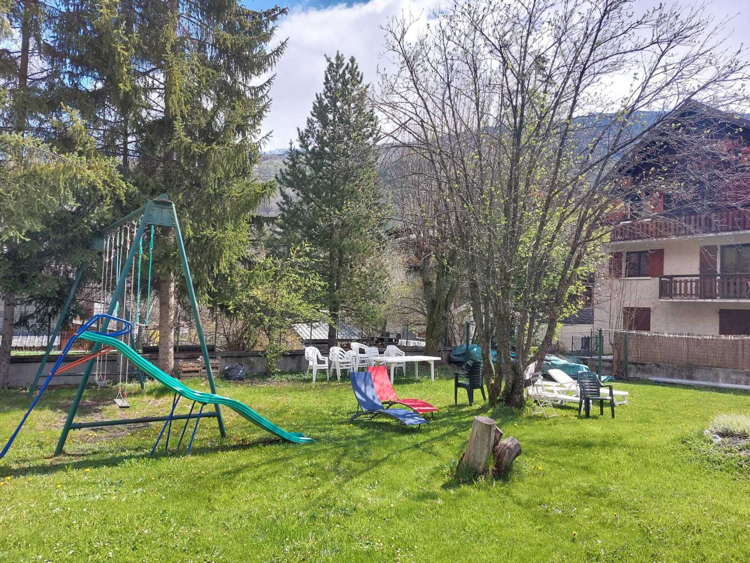 Ein Spielplatz mit Rutschen und Schaukeln, umgeben von Bäumen und einem Gebäude mit Balkonen.
