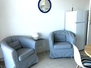 Una habitación con dos sillones, una mesita con lámpara, un refrigerador blanco y una mesa redonda con mantel a rayas.