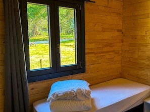 Una cama individual con colchón y almohadas blancos en una habitación con paredes de madera y ventana con vistas a un área verde.