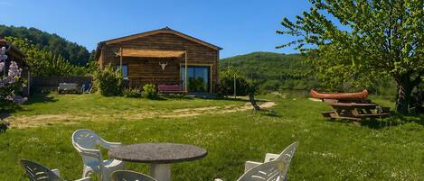 A wooden cabin with a porch, surrounded by a grassy yard and a tree.