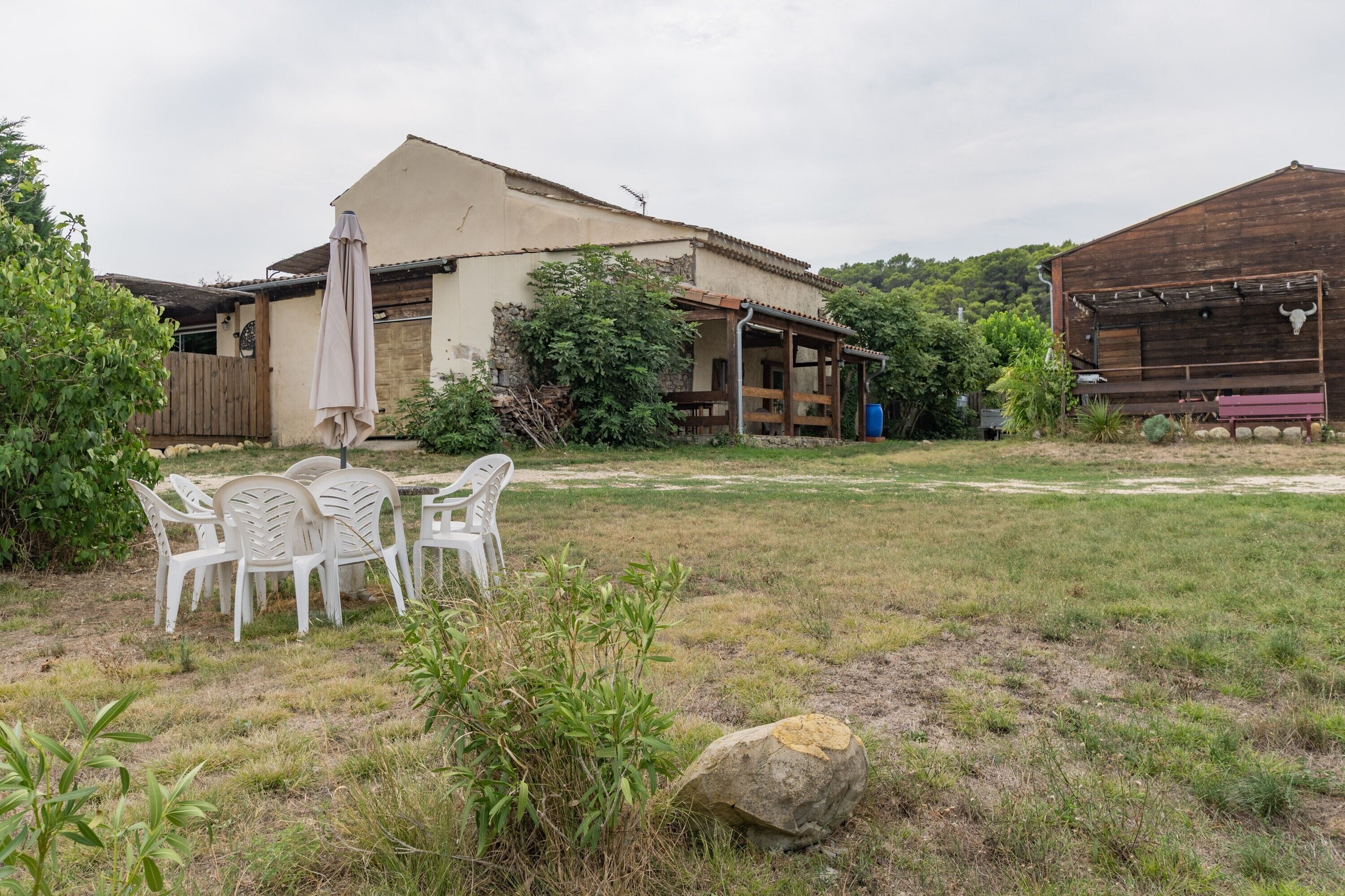Ein rustikaler Sitzbereich im Freien mit Plastikstühlen und einem Tisch, umgeben von einer Rasenfläche und einem Holzhaus mit Veranda.