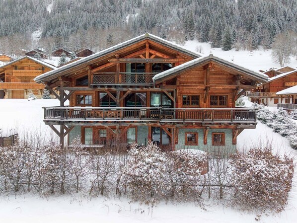 A multi-story wooden chalet with balconies, surrounded by snow-covered trees and a mountainous backdrop.