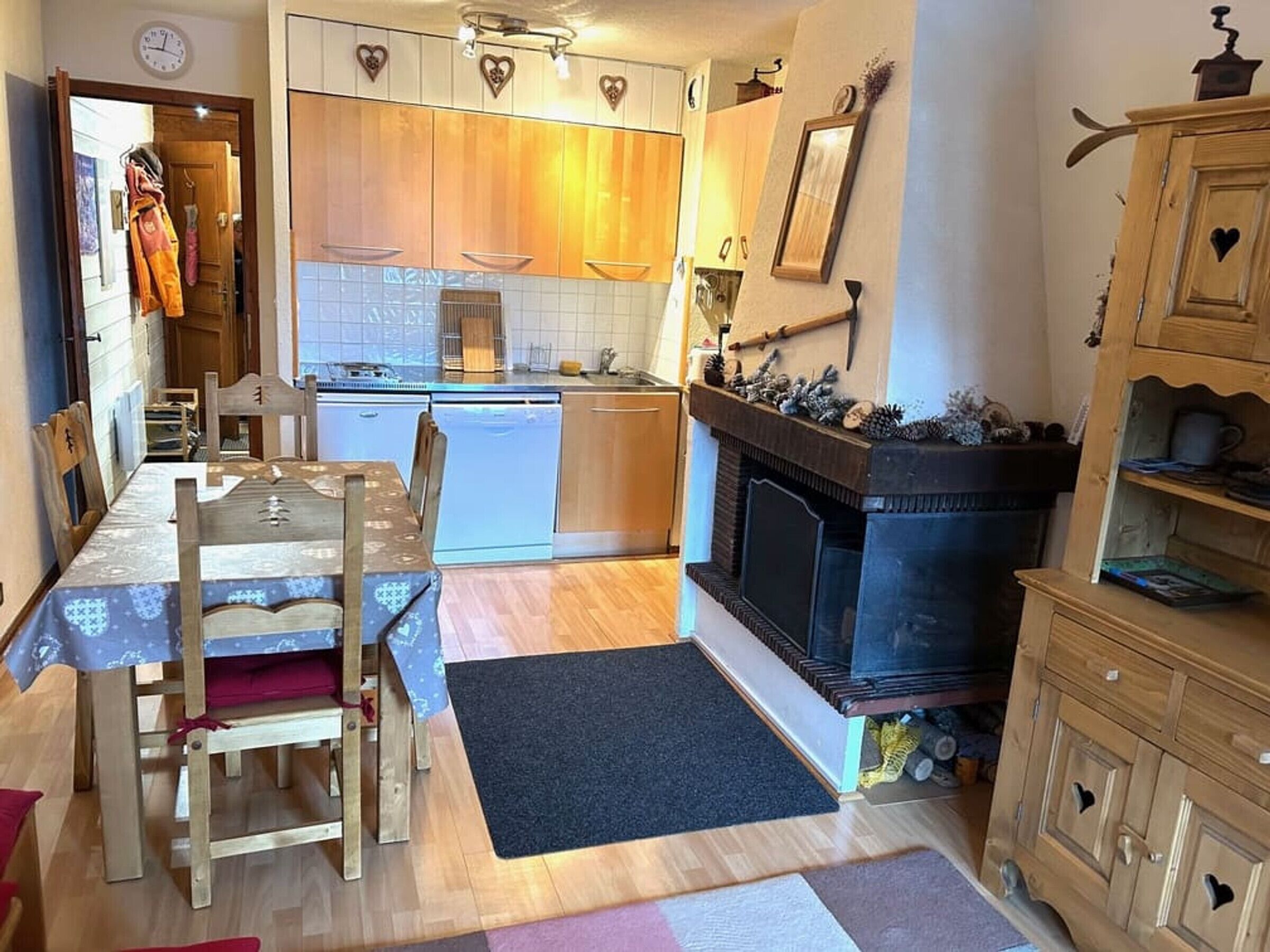 A compact kitchen with wooden cabinets, a dining table with chairs, a black fireplace, and a clock on the wall.