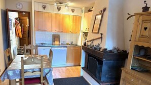 Une cuisine de taille réduite avec des meubles de rangement en bois, une table à manger avec des chaises, une cheminée noire et une horloge au mur.