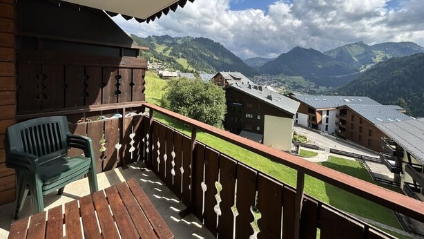 Un balcon en bois offrant une vue sur un village de montagne, une chaise en plastique verte et une table en bois.