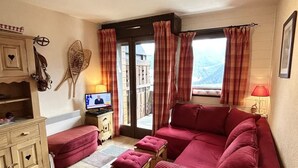 Un salon chaleureux avec un canapé rouge, un meuble en bois, une télévision et une vue sur l’extérieur par une fenêtre ornée de rideaux.