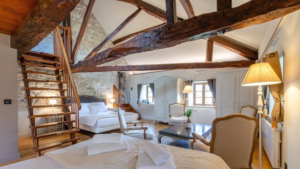 A bedroom with a wooden staircase, a bed with white linens, a chair, a lamp, and a view of a bathroom. - Guest Room – Château de la Rive – Sleeps 10 (Cruet)