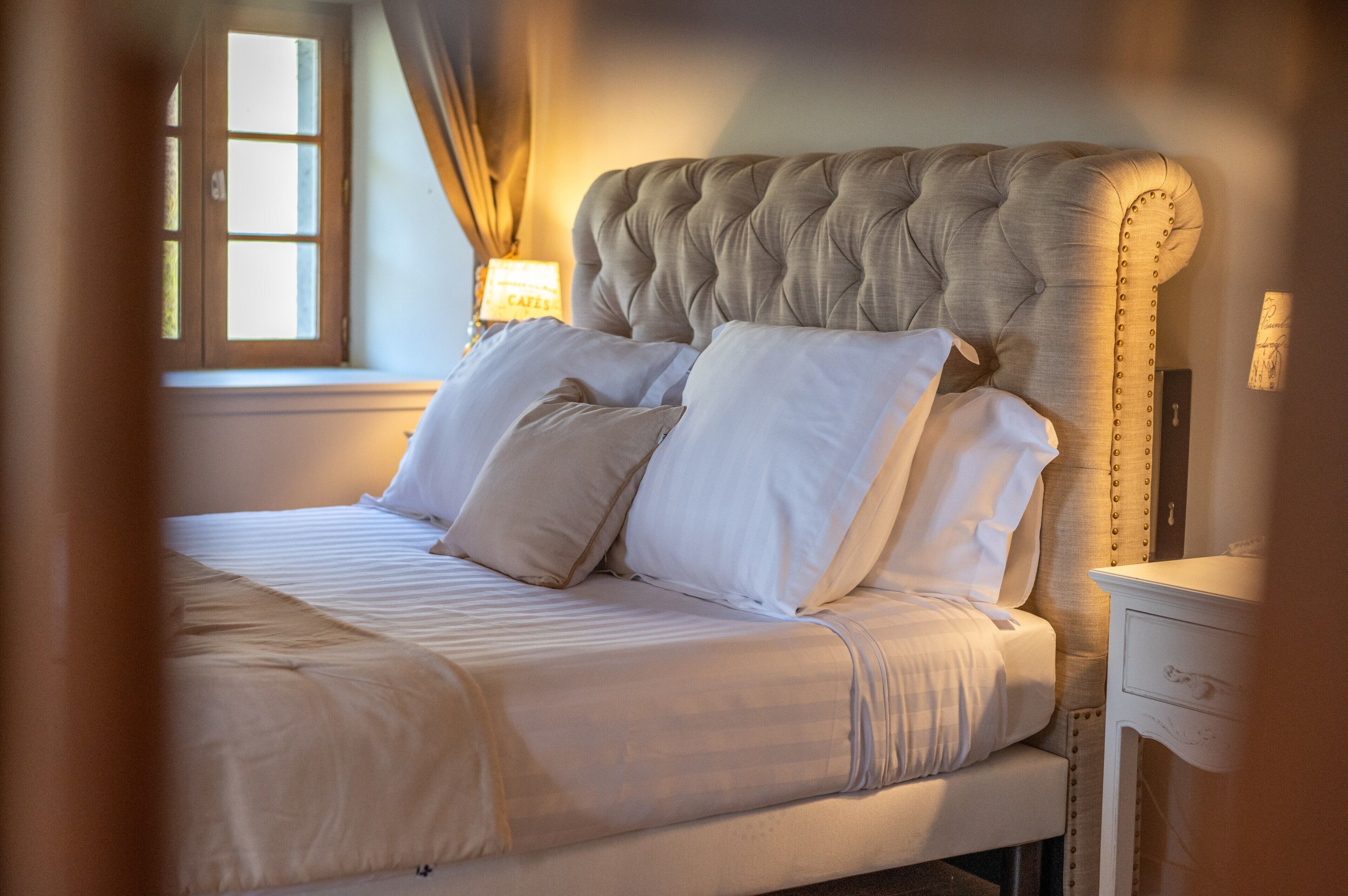A neatly made bed with a tufted headboard, a bedside lamp, and a window with curtains.