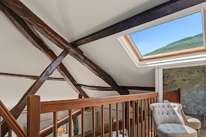 A room with a sloped ceiling, wooden beams, a window with a view of a green landscape, a wooden railing, and a tufted chair. - Guest Room – Château de la Rive – Sleeps 10 (Cruet)