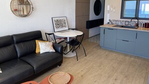 A modern living room with a black leather sofa, a round dining table, and a kitchen area with blue cabinets and a window overlooking the sea. - T2 Apartment with AC and Sea Views in Canet-en-Roussillon (Canet-en-Roussillon)