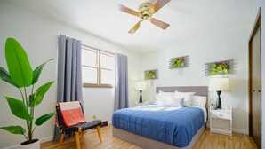 Une chambre à coucher comprenant un lit, une chaise, une plante en pot, un ventilateur de plafond et deux lampes fixées au mur.