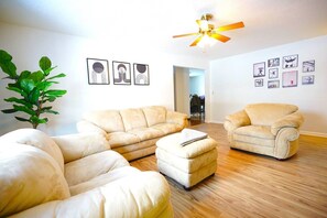 Ein Wohnzimmer mit einem beigefarbenen Ecksofa, einem passenden Sessel und einem kleinen Hocker. Es gibt einen Deckenventilator, einen Blumentopf und gerahmte Bilder an den Wänden.