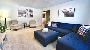 Sala de estar con un sofá seccional azul, dos sillones, una mesita auxiliar y una lámpara. Se observan cuadros enmarcados en la pared y un cuadro grande de un toro.