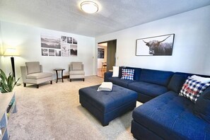 A living room with a blue sectional sofa, two armchairs, a small side table, and a lamp. There are framed pictures on the wall and a large bull painting. - Grand Duplex Blue House (Sioux Falls)