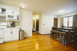 A compact kitchen with white cabinets, a microwave, and a coffee maker. There is a dining area with a wooden table and chairs.