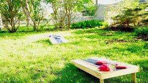 Une table en bois, sur laquelle sont posés un coussin rouge et un objet noir, est placée sur une pelouse verdoyante. On aperçoit des arbres et une clôture en arrière-plan.